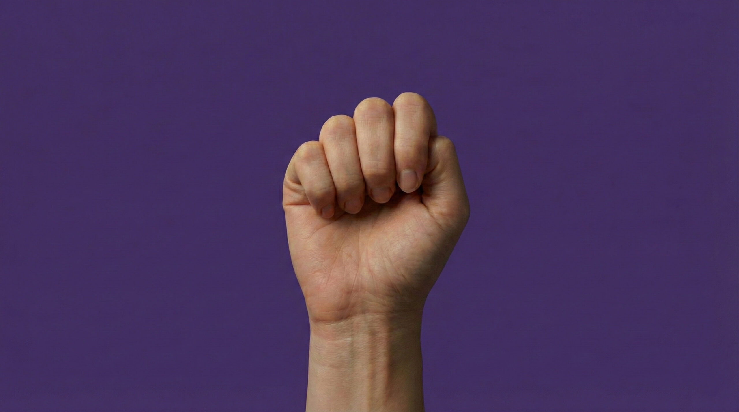 Mano levantada en forma de puño sobre un fondo morado, símbolo de reivindicación y fuerza en el Día Internacional de la Eliminación de la Violencia contra la Mujer por el símbolo de ayuda en casos de violencia de género.